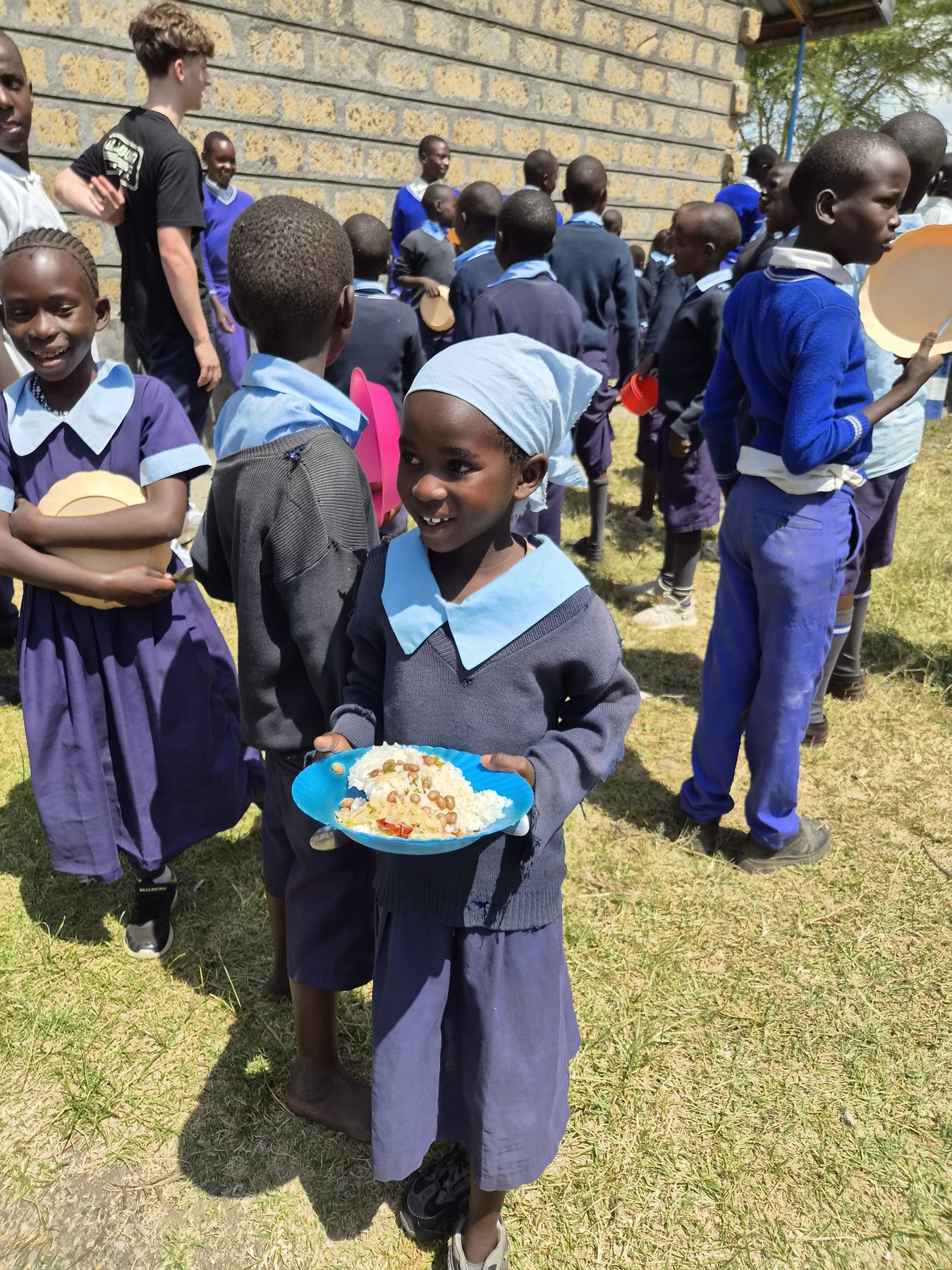 A primary school student holding a plate of lunch with classmates nearby